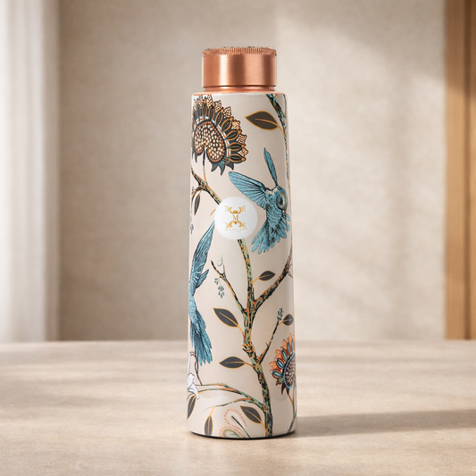 A tall insulated water bottle with a cream background and a detailed botanical pattern featuring bluebirds, sunflowers, and branches. The bottle has a metallic rose gold cap and is standing on a light beige surface against a softly lit, neutral background.